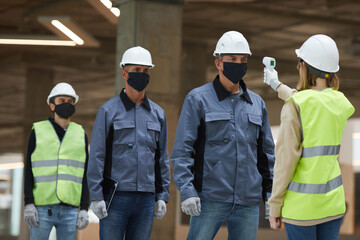 Portrait of female supervisor measuring temperature of workers with contactless thermometer at construction site, corona virus safety measures
