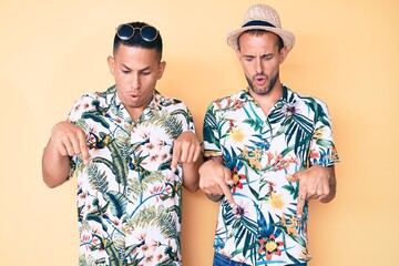 Young gay couple of two men wearing summer hat and hawaiian shirt pointing down with fingers showing advertisement, surprised face and open mouth