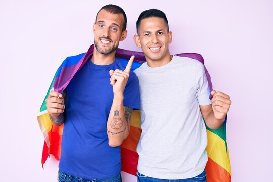 Young gay couple of two men holding rainbow lgbtq flag together smiling happy pointing with hand and finger to the side