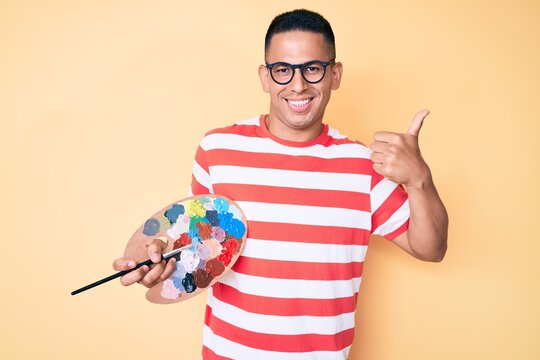 Young handsome latin man holding paintbrush and palette smiling happy and positive, thumb up doing excellent and approval sign