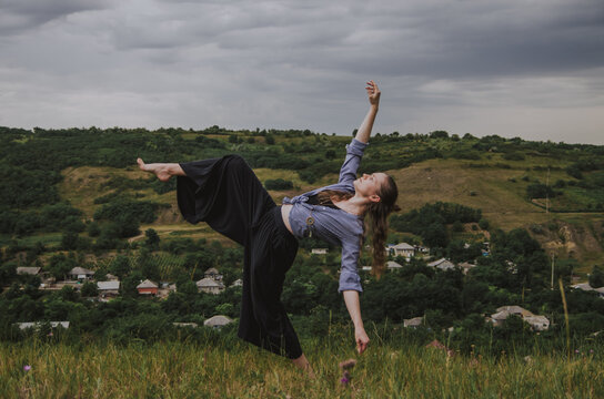 Woman Wearing Large Pants And Striped Shirt Dancing With One Leg Up And Hands In The Air On A Hill In A Rural Setting. Concept: Self Expression, Freedom Of Movement, Dance