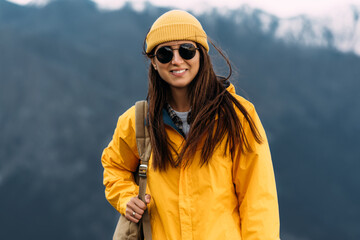Fototapeta na wymiar A young woman in Hiking gear stands against the backdrop of mountains and looks at the camera. A woman among the mountains. Traveling woman with a backpack on the background of mountains. Copy space