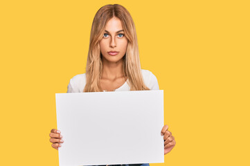Naklejka premium Beautiful blonde young woman holding blank empty banner thinking attitude and sober expression looking self confident