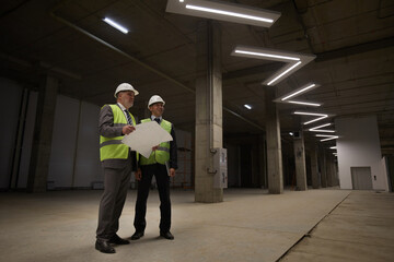 Low angle view at two business people wearing hardhats and holding plans while standing at construction site, copy space