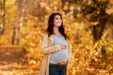 Attractive millennial pregnant woman holding her tummy in autumn park