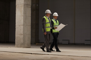 Full length portrait of two business people wearing hardhats and holding plans while walking at construction site indoors, copy space - obrazy, fototapety, plakaty