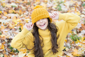 happy teenage kid having fun in autumn forest with beautiful seasonal maple leaves wearing warm knitted clothes such as hat and sweater, autumn fun