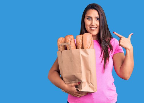 Young beautiful brunette woman holding delivery bag with bread pointing finger to one self smiling happy and proud