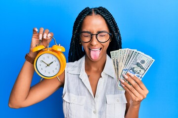 Beautiful hispanic woman holding alarm clock and dollars sticking tongue out happy with funny...