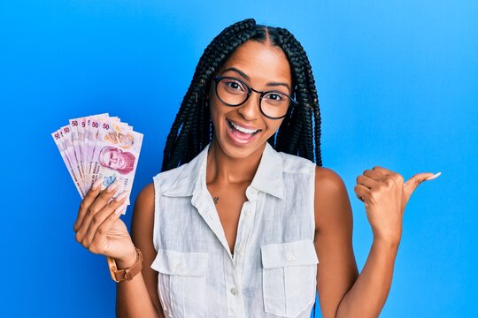Beautiful hispanic woman holding 50 mexican pesos banknotes pointing thumb up to the side smiling happy with open mouth