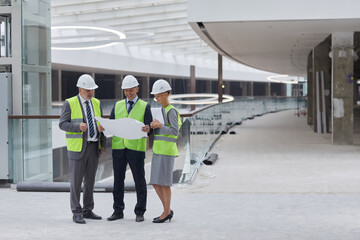 Full length portrait of three successful business people wearing hardhats and inspecting plans...