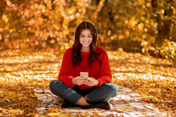 Pretty young woman sitting on picnic blanket at autumn park with headphones and mobile device and listening to music