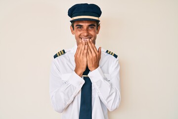 Young hispanic man wearing airplane pilot uniform laughing and embarrassed giggle covering mouth...