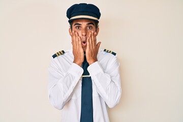 Young hispanic man wearing airplane pilot uniform afraid and shocked, surprise and amazed expression with hands on face