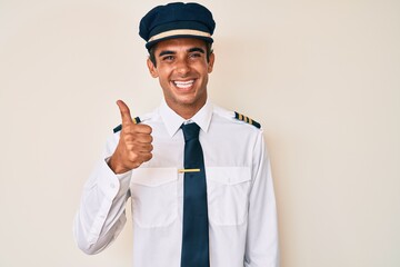 Young hispanic man wearing airplane pilot uniform doing happy thumbs up gesture with hand. approving expression looking at the camera showing success.