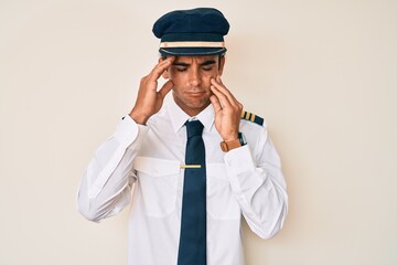 Young hispanic man wearing airplane pilot uniform with hand on head, headache because stress. suffering migraine.