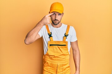 Young handsome man wearing handyman uniform over yellow background pointing unhappy to pimple on forehead, ugly infection of blackhead. acne and skin problem
