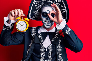 Young man wearing mexican day of the dead costume holding alarm clock smiling happy doing ok sign with hand on eye looking through fingers