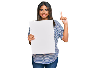 Young latin girl holding blank empty banner surprised with an idea or question pointing finger with happy face, number one