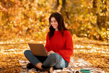 Lovely Caucasian woman using laptop for online work, browsing web at city park in autumn