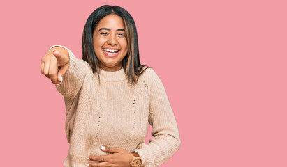 Young latin girl wearing wool winter sweater laughing at you, pointing finger to the camera with hand over body, shame expression