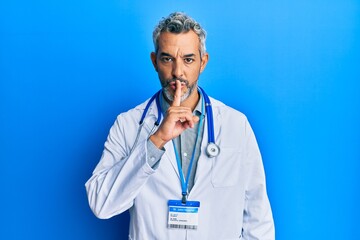 Middle age grey-haired man wearing doctor uniform and stethoscope asking to be quiet with finger on...