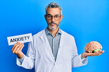 Middle age grey-haired man wearing doctor coat holding brain and anxiety message relaxed with serious expression on face. simple and natural looking at the camera.