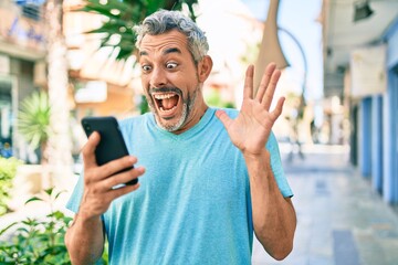 Middle age grey-haired man using smartphone at street of city celebrating victory with happy smile and winner expression with raised hands