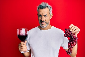 Middle age grey-haired man holding branch of fresh grapes and red wine clueless and confused expression. doubt concept.