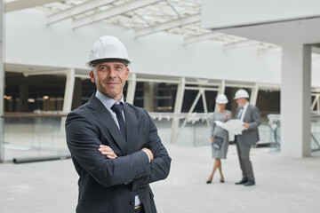 Waist up portrait of smiling mature businessman wearing hardhat and looking at camera while...