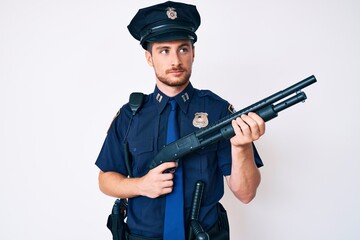 Young caucasian man wearing police uniform holding shotgun smiling looking to the side and staring away thinking.