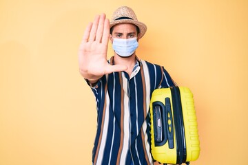 Young handsome man wearing medical mask holding suitcase with open hand doing stop sign with serious and confident expression, defense gesture