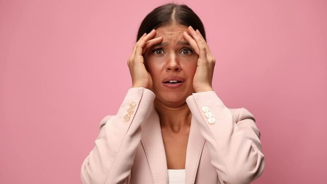 unhappy woman in pink suit covering face with hands, making scared faces, playing and grimacing on pink background
