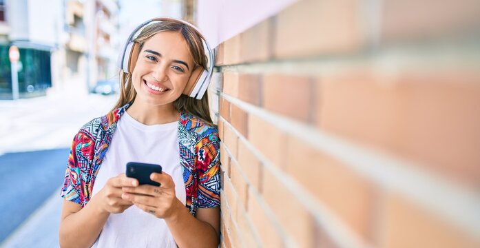Young beautiful blonde caucasian woman smiling happy outdoors on a sunny day wearing headphones and using smartphone leaning on a brick wall
