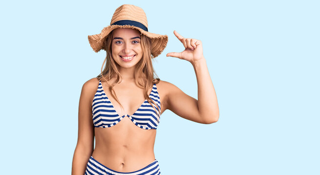 Young beautiful blonde woman wearing bikini and hat smiling and confident gesturing with hand doing small size sign with fingers looking and the camera. measure concept.