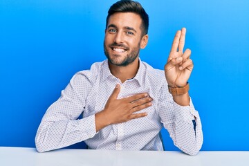 Handsome hispanic man wearing business clothes sitting on the table smiling swearing with hand on...