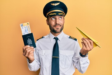 Handsome hispanic pilot man holding paper plane and passport smiling looking to the side and staring away thinking. © Krakenimages.com