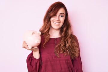 Young beautiful woman holding piggy bank looking positive and happy standing and smiling with a confident smile showing teeth