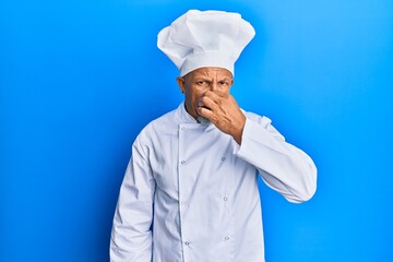 Middle age grey-haired man wearing professional cook uniform and hat smelling something stinky and disgusting, intolerable smell, holding breath with fingers on nose. bad smell