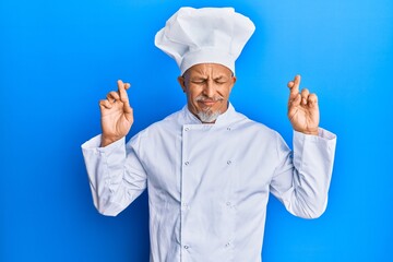 Middle age grey-haired man wearing professional cook uniform and hat gesturing finger crossed smiling with hope and eyes closed. luck and superstitious concept.