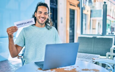 Young hispanic man smiling happy working using laptop and holding mindfulness word paper at terrace of coffee shop.