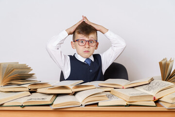 Education concept. The student does his homework, holds his head with his hands and looks at the camera. There are many open books on the table.