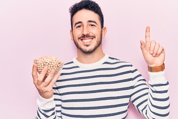 Young hispanic man holding chickpeas bowl smiling with an idea or question pointing finger with happy face, number one