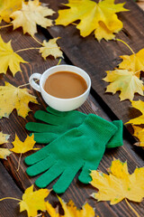 Cup of coffee and green gloves next to maple leaves on wooden table.