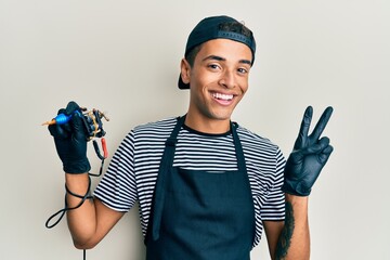 Young handsome african american man tattoo artist wearing professional uniform and gloves holding tattooer machine smiling looking to the camera showing fingers doing victory sign. number two.
