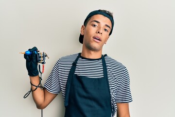 Young handsome african american man tattoo artist wearing professional uniform and gloves holding tattooer machine looking sleepy and tired, exhausted for fatigue and hangover.