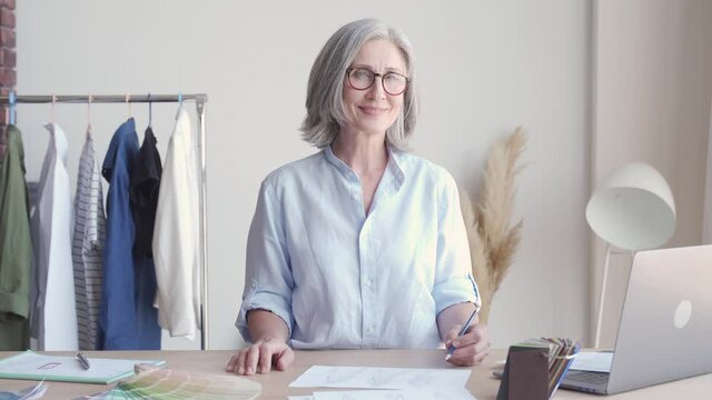 Happy middle aged stylish woman fashion designer wears glasses standing at workplace desk, portrait. Smiling sophisticated 60s old lady dressmaker drawing sketches, looking at camera in modern atelier