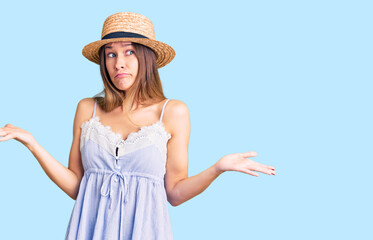 Beautiful brunette young woman wearing summer hat clueless and confused expression with arms and hands raised. doubt concept.