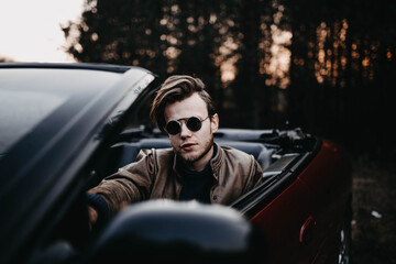  brutal man with a beard sits in a red convertible in sunglasses in nature. Male bachelor and driver style.