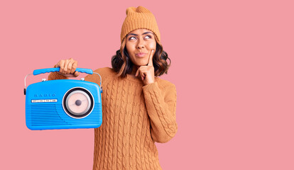 Young beautiful mixed race woman holding vintage radio serious face thinking about question with...